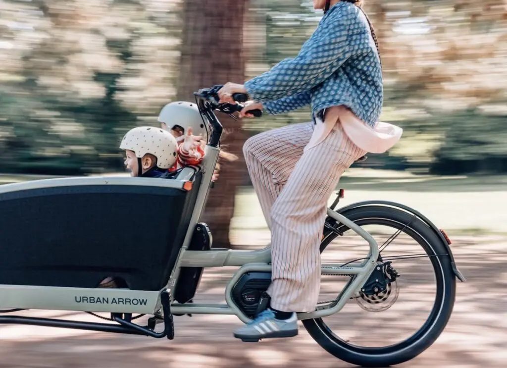 Parent riding an Urban Arrow cargo bike with two children wearing helmets in a park-like setting, blurred background due to motion.