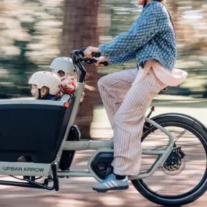 Parent riding an Urban Arrow cargo bike with two children wearing helmets in a park-like setting, blurred background due to motion.