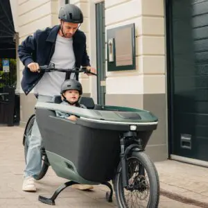 Adult man pushes a cargo bike with a child wearing a helmet in the front passenger box along a city sidewalk.