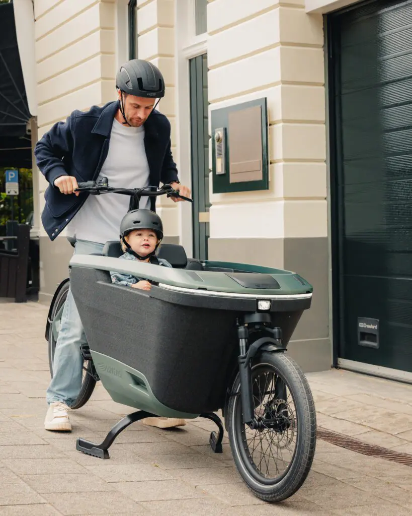 Adult man pushes a cargo bike with a child wearing a helmet in the front passenger box along a city sidewalk.