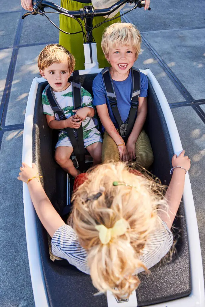 Two smiling young children strapped into a white bike trailer with safety harnesses, attached to a bicycle.
