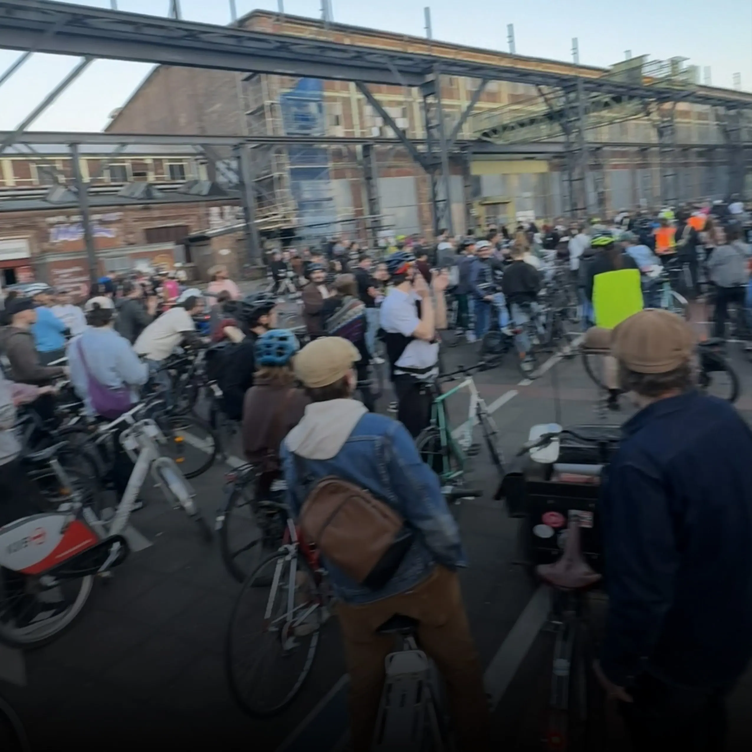 Crowded city street with many cyclists and bystanders beneath a metal overhead structure, urban buildings in the background