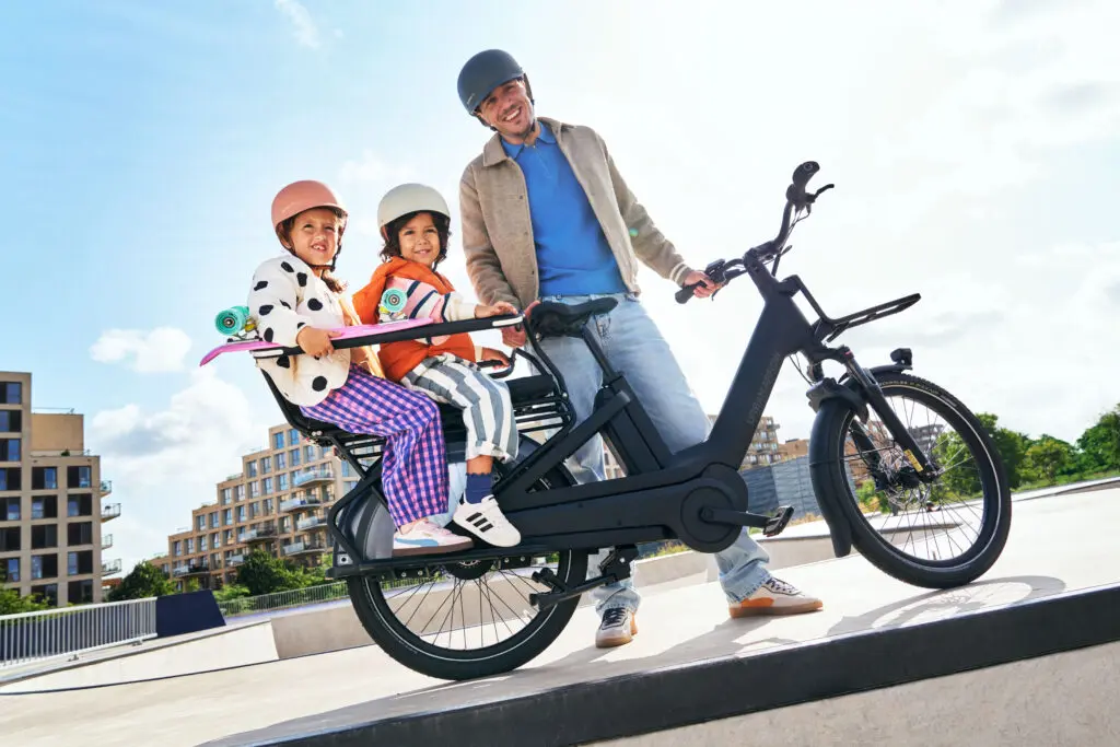 Father and two children on an electric bike wearing helmets, riding through a sunny urban area.