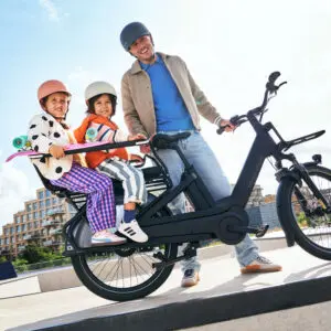 Father and two children on an electric bike wearing helmets, riding through a sunny urban area.