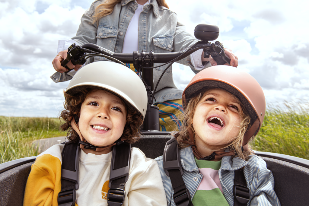 Three children with helmets smile at the camera while riding a bike outdoors on a sunny day.