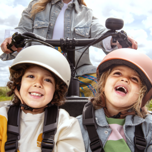 Three children wearing helmets smile at the camera while riding a bike outdoors on a sunny day.