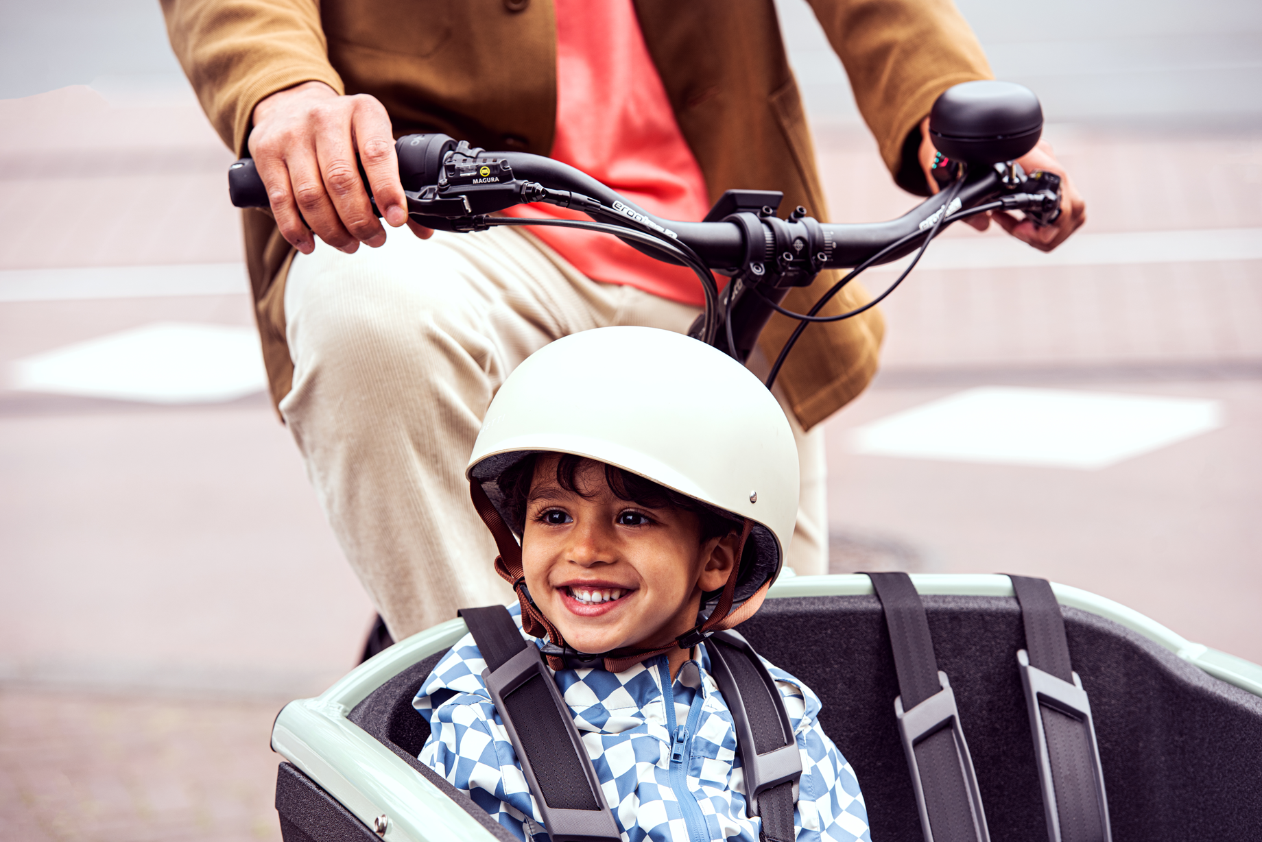 Urban_Arrow_FamilyNext_Green_592 - mycargobike köln GmbH Smiling young boy wearing a white bike helmet sits in a child bike seat, with an adult guiding the handlebars behind him.