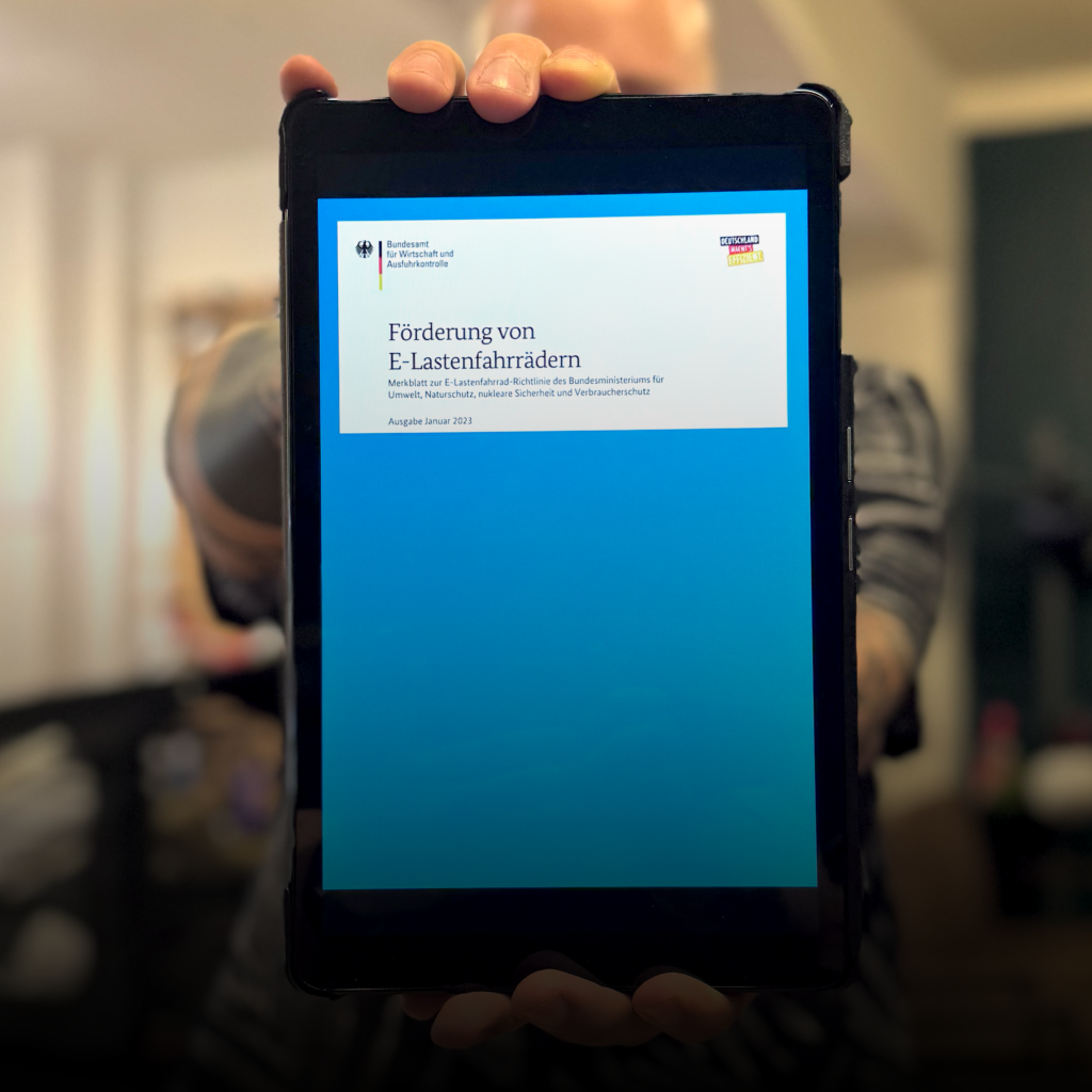 Close-up of a hand holding a tablet in a black case, showing a German document titled 'Förderung von E-Lastenfahrrädern'.