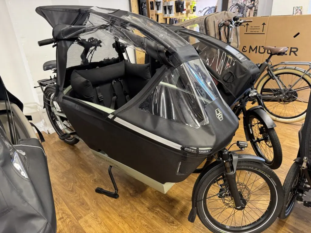 Black cargo bike with a large front-loading box covered in clear plastic, indoors in a showroom setting.