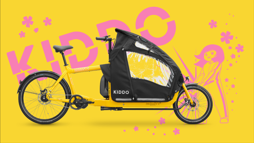 Yellow cargo bike with a large black canopy and Kiddo branding, set against a bright yellow background with pink floral and graphic accents.