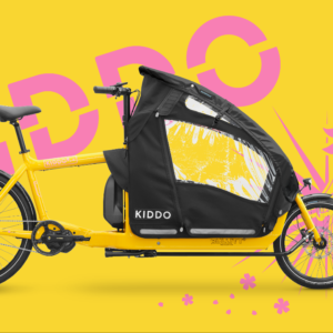 Yellow cargo bike with a large black canopy and Kiddo branding, set against a bright yellow background with pink floral and graphic accents.
